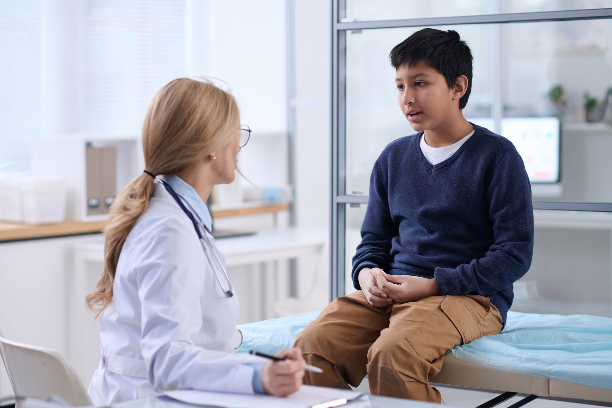 A young boy sits on exam table speaking to a doctor.