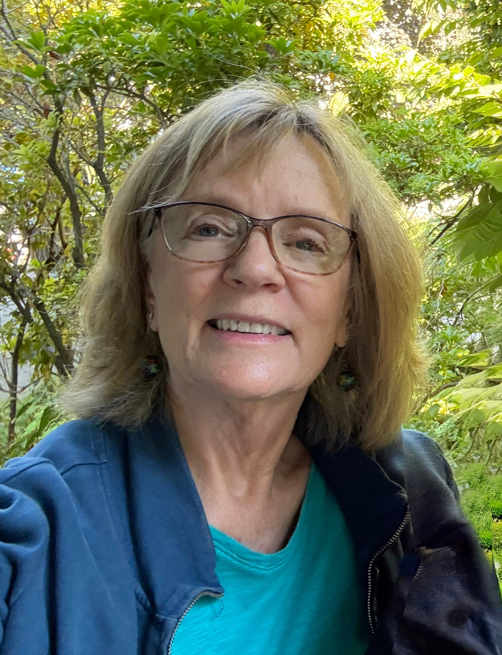 Headshot of woman with short, light brown hair and glasses, wearing a blue blazer over a teal shirt, with trees in the background