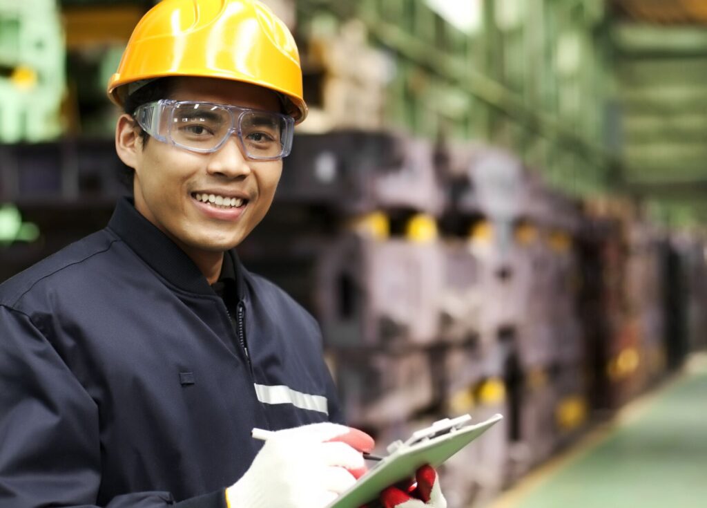 Warehouse worker with dark blue jacket, yellow hardhat, safety glasses, clipboard