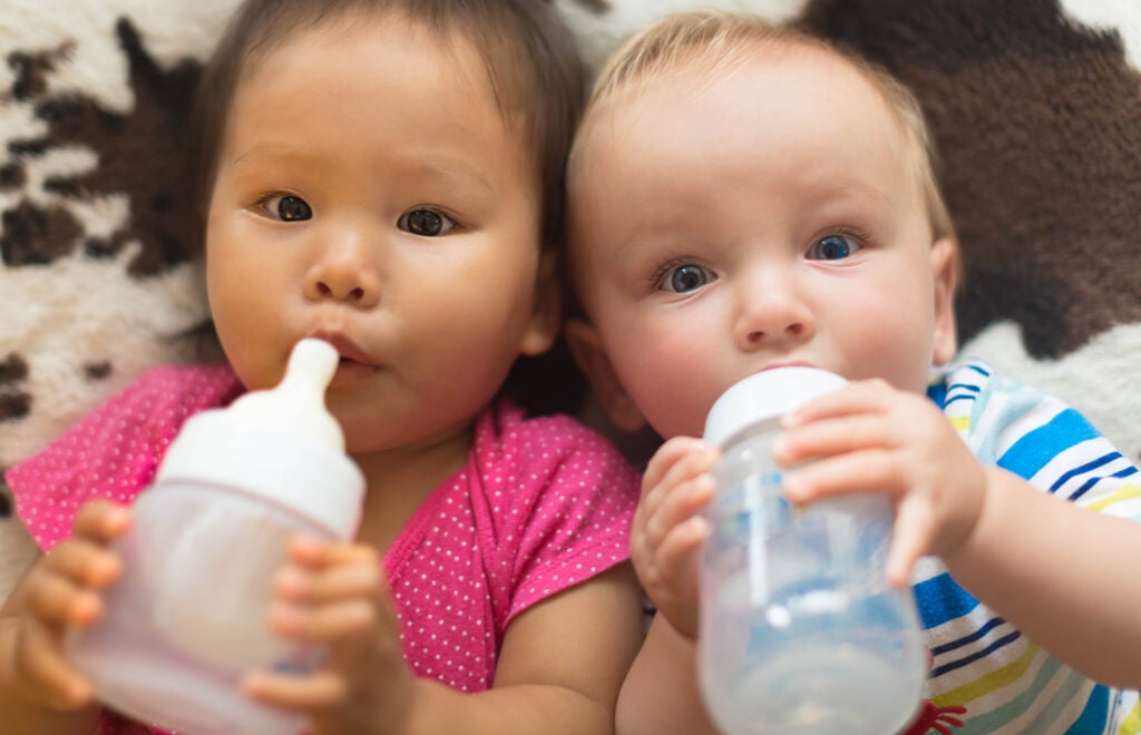 Asian and Caucasian babies laying on the floor side by side drinking from baby bottles
