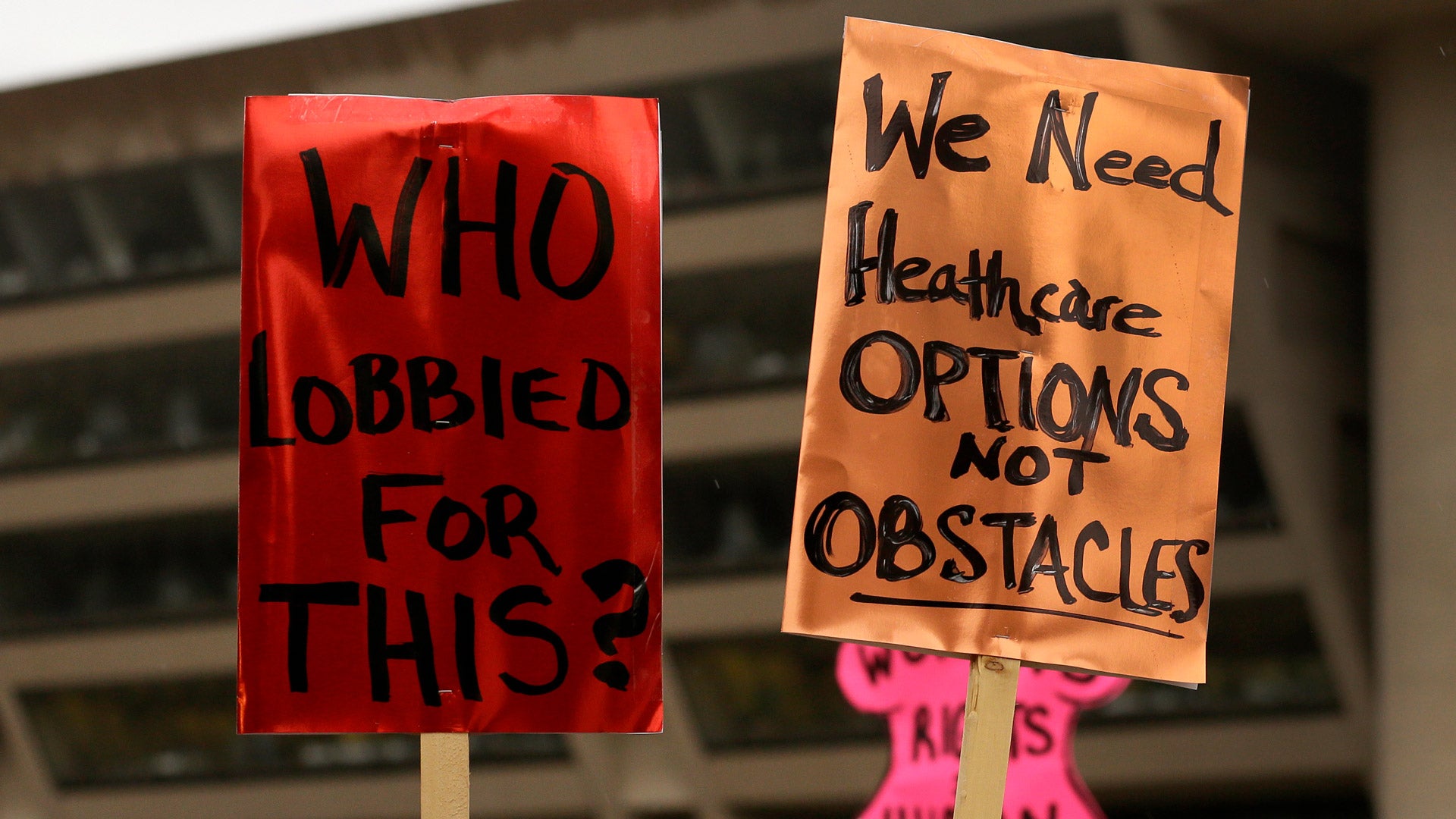 A red protest sign (left) and an orange protest sign (right) are held in the air. The red one reads “Who lobbied for this?” in black text. The orange one reads “We need healthcare options not obstacles.”
