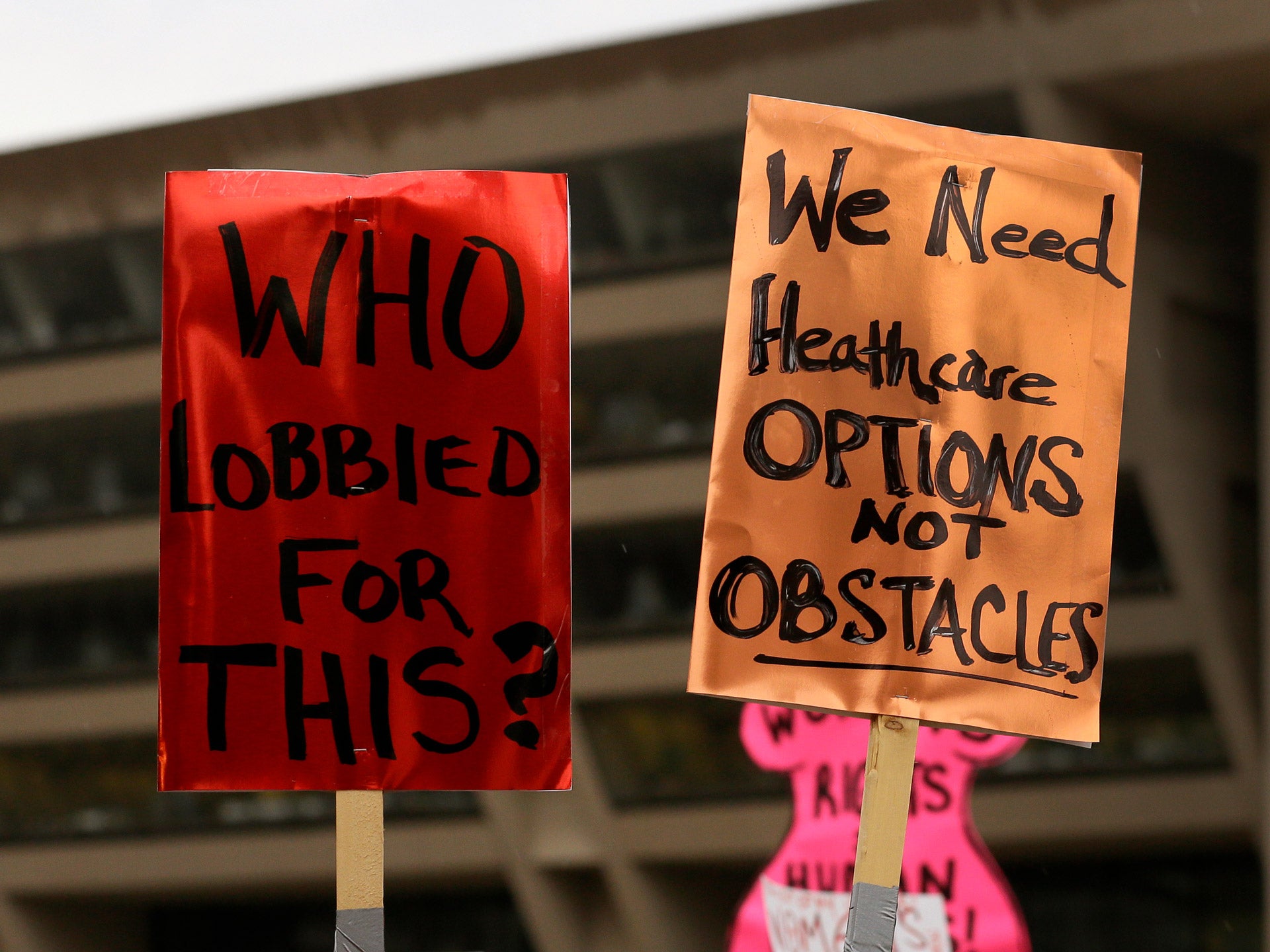 A red protest sign (left) and an orange protest sign (right) are held in the air. The red one reads “Who lobbied for this?” in black text. The orange one reads “We need healthcare options not obstacles.”