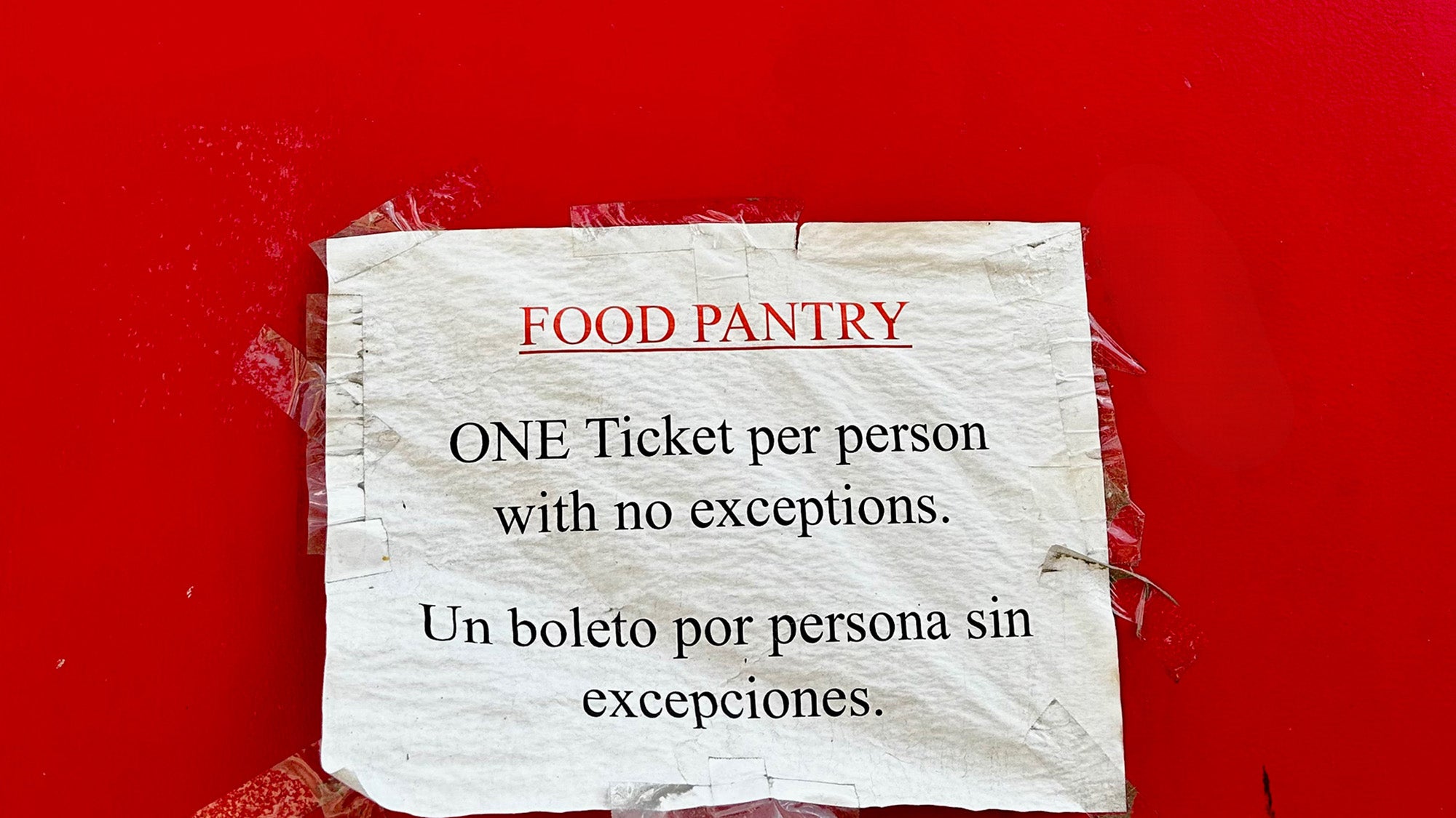 Typewritten instructions for accessing a Food Pantry for free food is detailed in an English and a Spanish note taped to a bright red back entry door at a Hoboken, New Jersey senior community center. The message states: “FOOD PANTRY ONE Ticket per person with no exceptions. Un boleto por persona sin excepciones.”