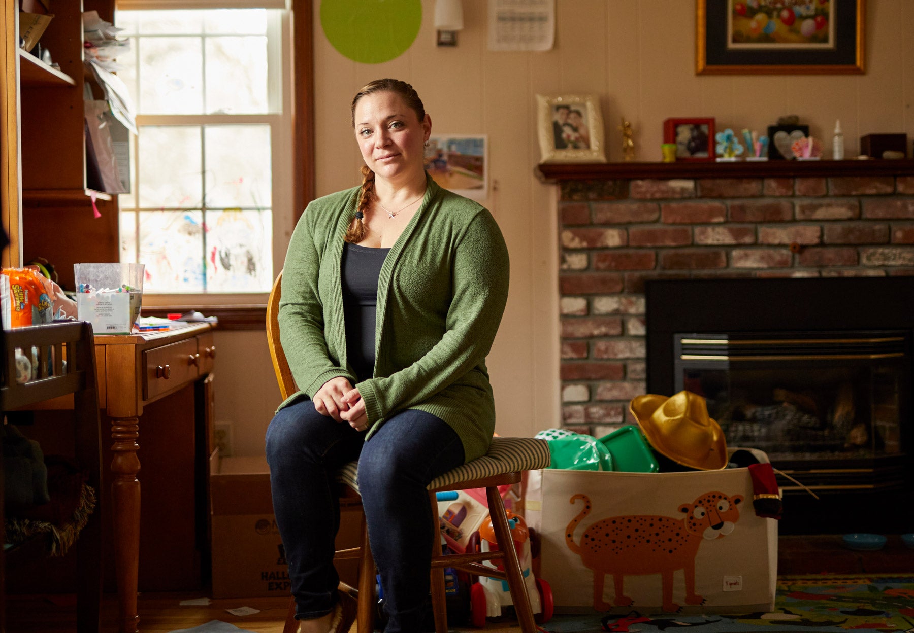 A white middle-aged woman sits in a living room. She wears a green cardigan, tank top and jeans, and her hair is braided in a single braid; hands clapsed in her lap. Surrounding her are children's toys, a bin on the floor and a fireplace with photos.