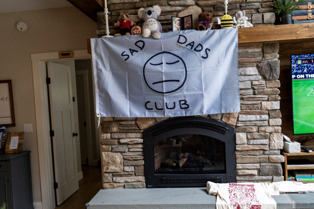 A white flag with the words "Sad dads club" hangs from a wooden mantle on a stone fireplace. Stuffed animals and two framed photo of an infant are on the mantle shelf. A TV with a sports game is on the right.