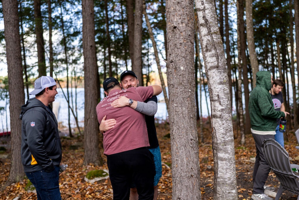 Two middle-aged men embrace in a pine-tree forest near a lake. Other men gather and converse nearby.