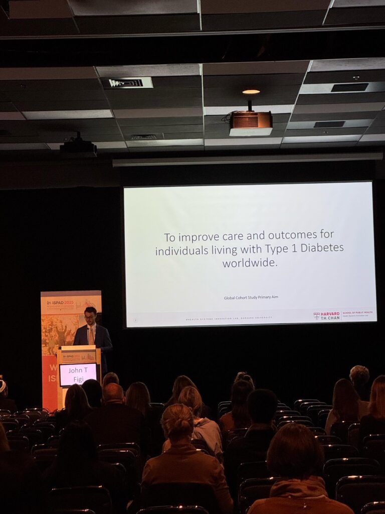 Jake Figi stands in front of an auditorium, presenting in front of a large screen.