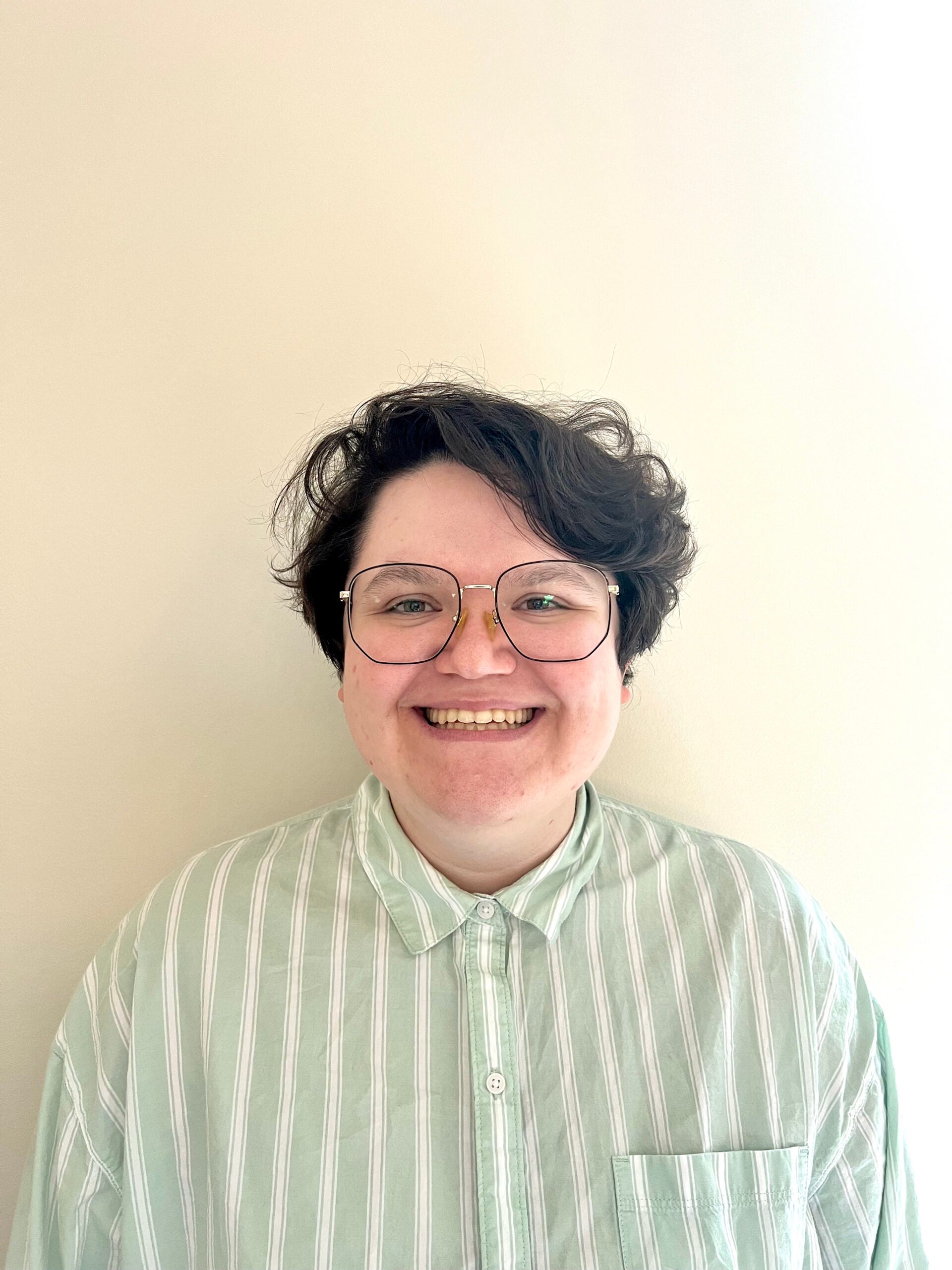 a headshot of Catherine Adcock. she wears glasses and has short, dark brown hair that parts from the viewer's left. she is smiling at the camera and wears a button-down, collared shirt that has one chest pocket and is green and white stripes. the background is white