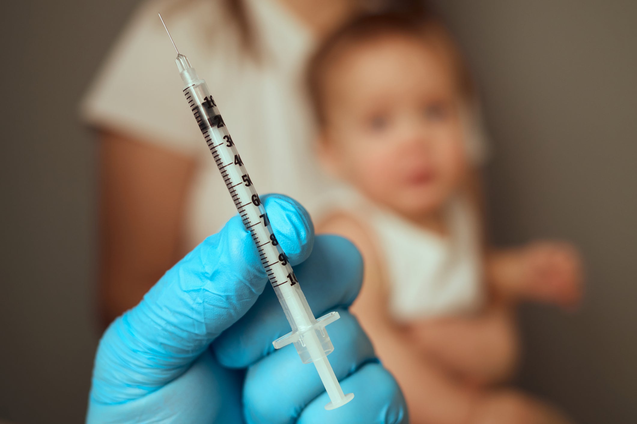 A gloved hand holds a syringe--representing a vaccine--in the foreground, with a mother holding a child in the background, out of focus.