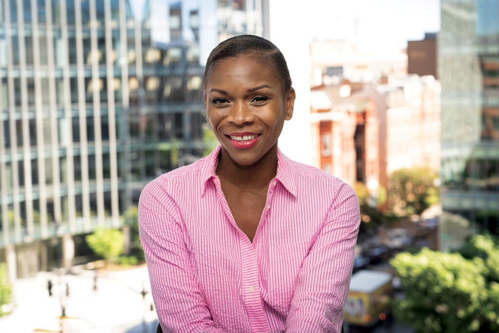Portrait of Abby Charles in front of an office window with the city in the background. She wears a pink and white button down shirt.