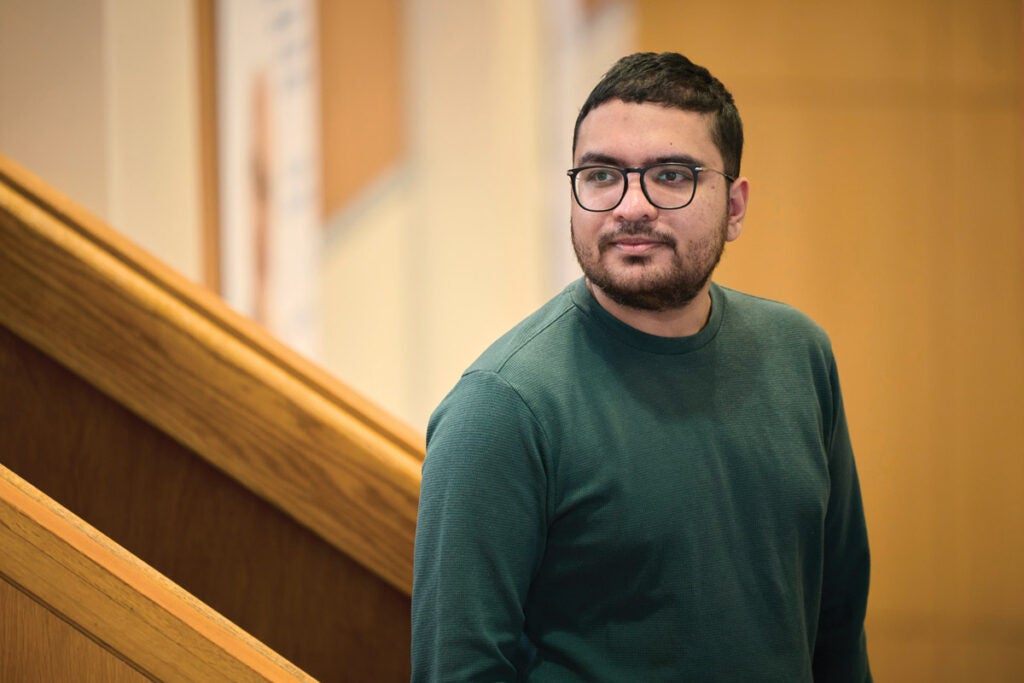 Akshay Naryanan stands for an portrait near a wooden railing. He wears a green sweater and glasses. 