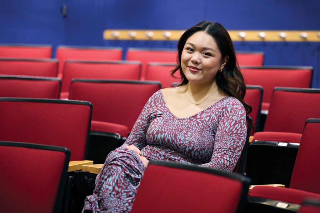 Carisha Prantoyo sits for a portrait while seated in an auditorium style classroom with red chairs. She wears a blue and purple patterned dress. 