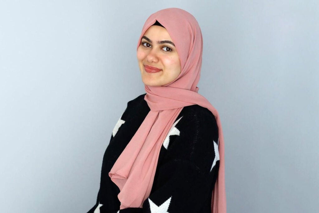 Nour Hammad stands for a portrait in front of a gray background. She wears a light pink hijab and a black sweater with white stars. 