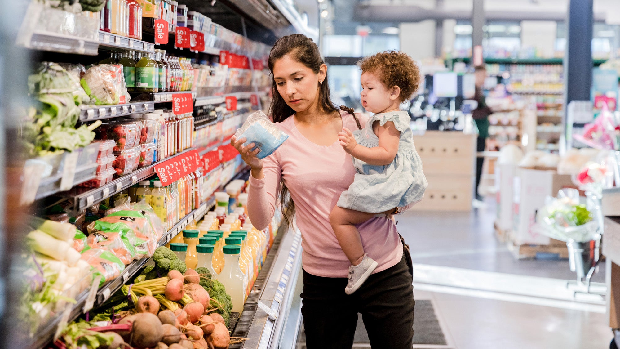 A young mother holds her daughter on her hip as she grocery shops for items she needs.