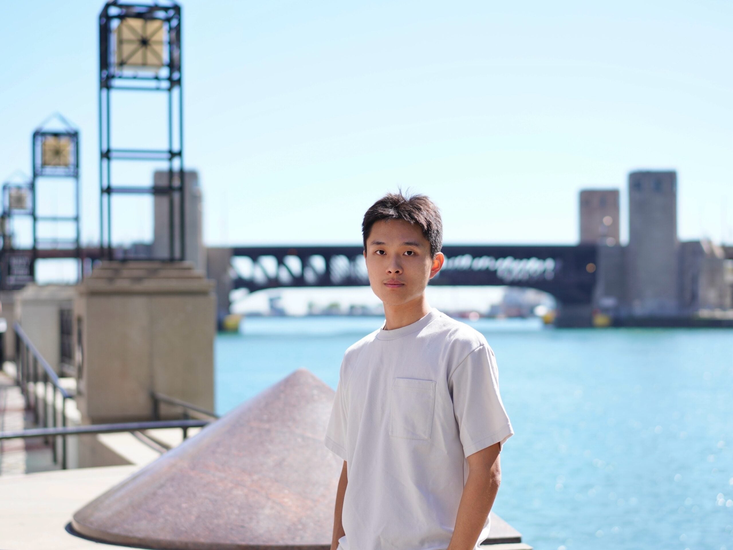 a headshot of Dr. Yu Gui. He looks directly at the camera and is wearing a white, short-sleeved shirt. the background is not in focus but shows a bridge over blue water and a light blue sky above