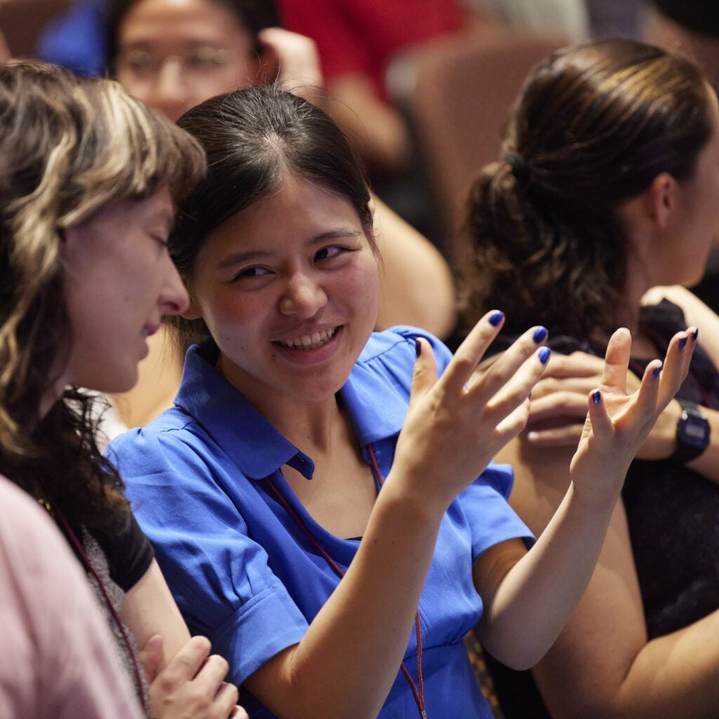 Two students chat in a full lecture hall, one with painted blue nails that match her top.