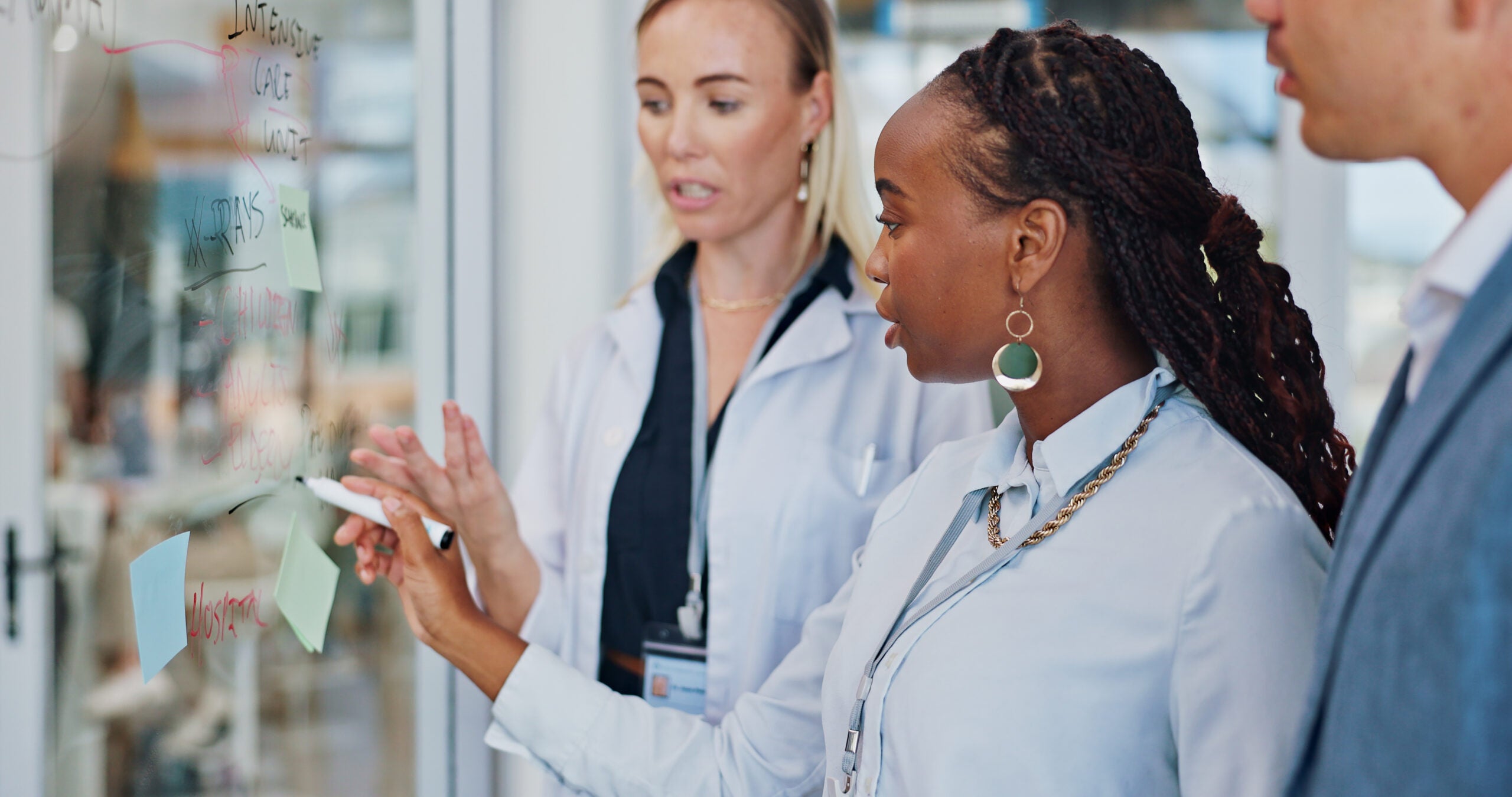 Female doctor writes on clear whiteboard with colleagues.