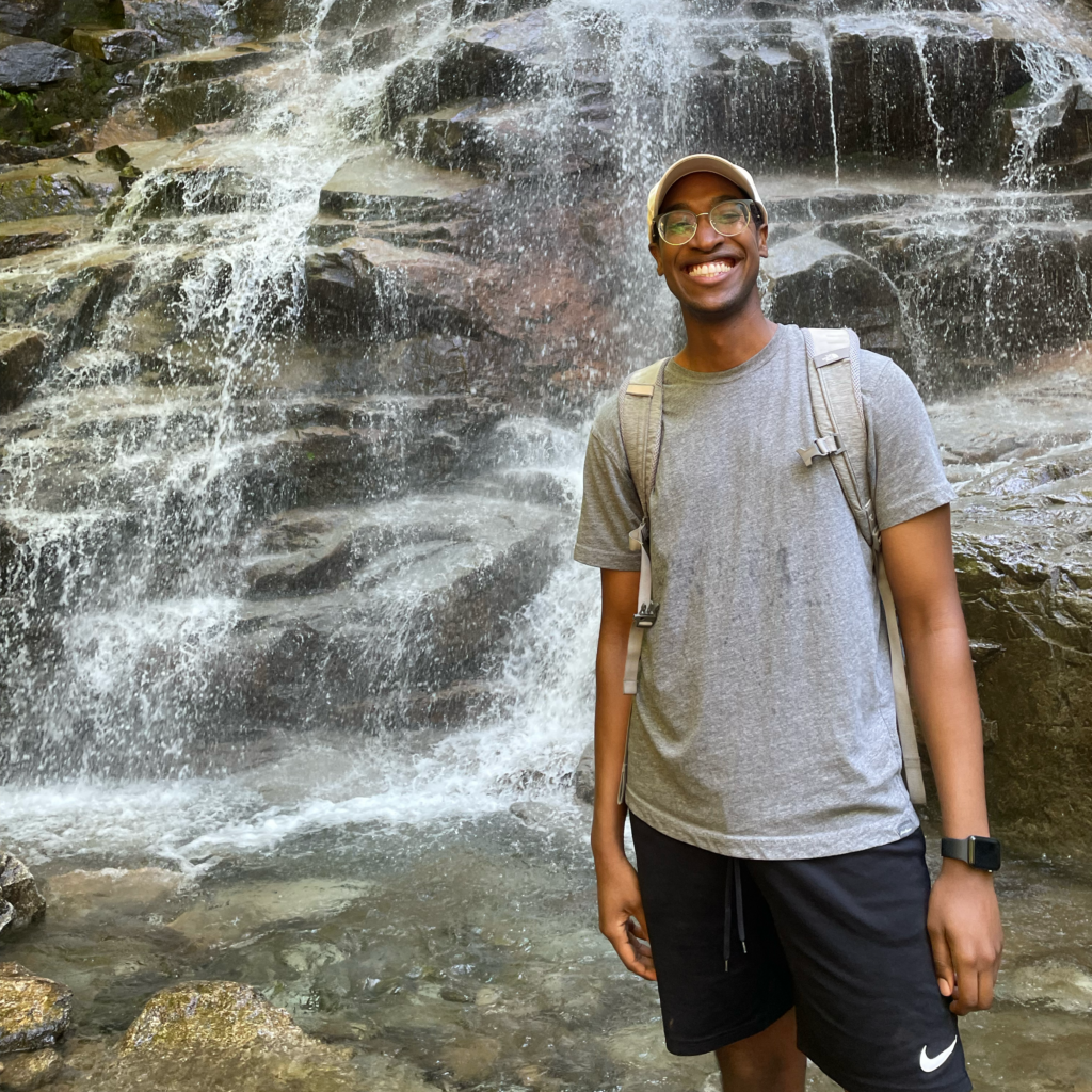 Samuel Zinga standing and smiling in front of a rocky waterfall, with clear water cascading down behind him into a shallow pool