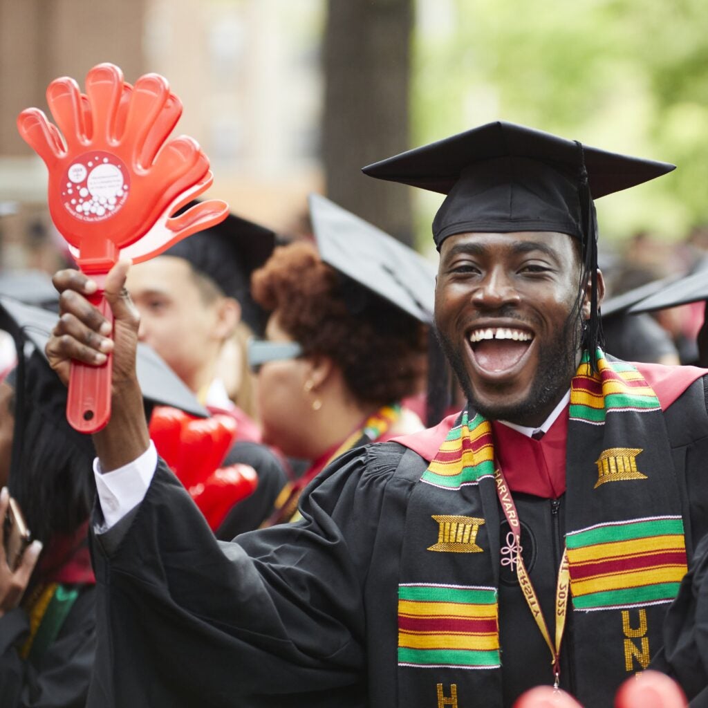 An exuberant graduate wearing a kente-inspired Black affinity stole shakes his Harvard Chan hand clapper. 