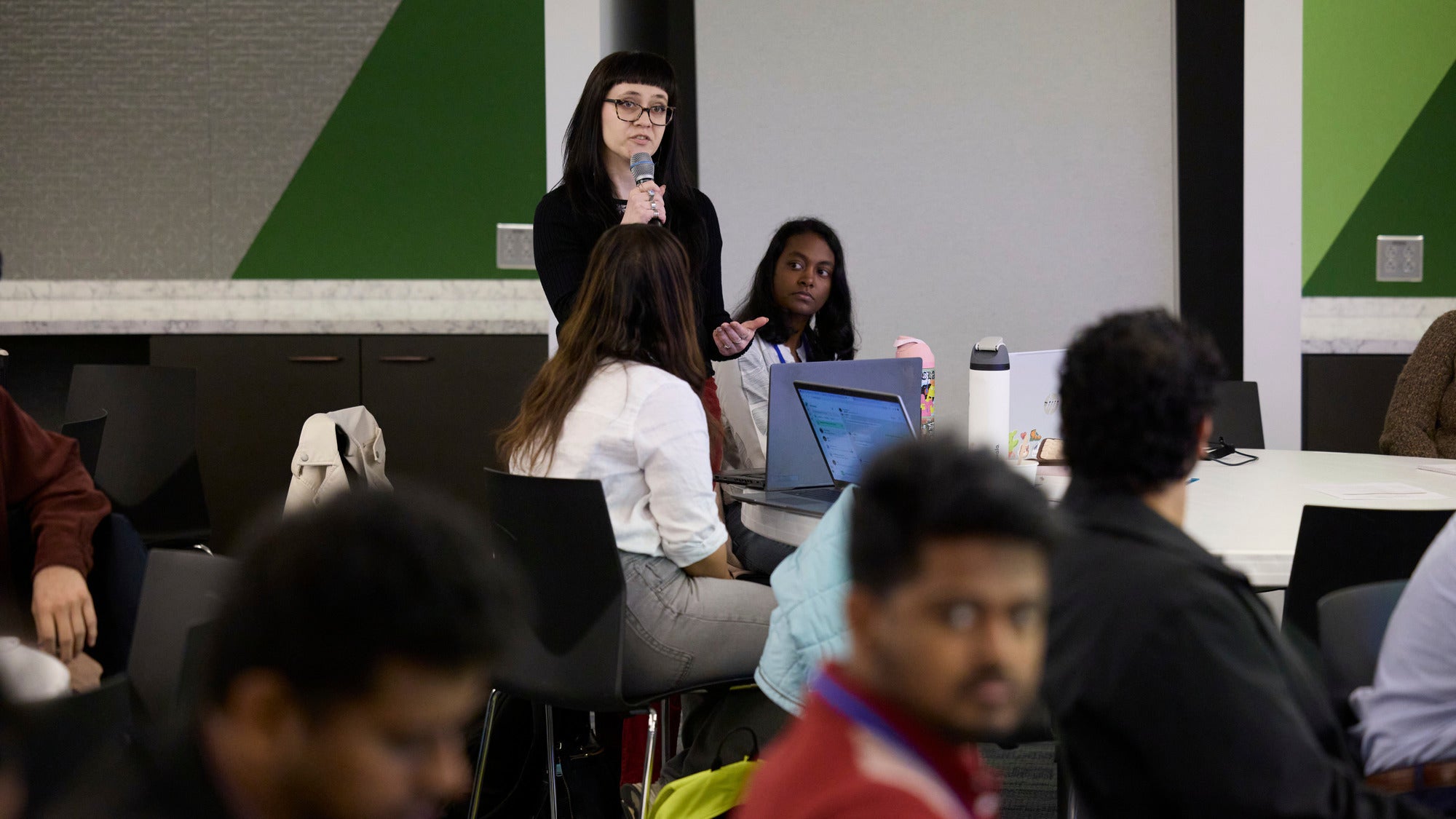 A standing woman speaks into a microphone as people sitting at tables look on