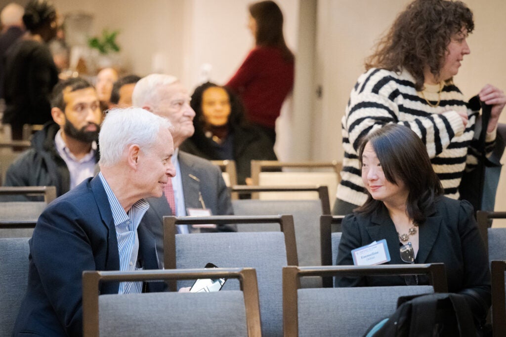 Two audience members lean across an empty seat to chat before the program starts. 