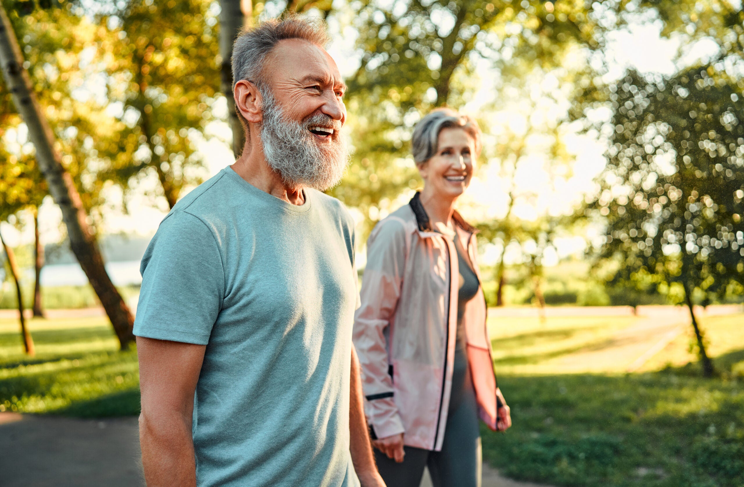Grey haired man and woman strolling at summer park and smiling cheerfully.