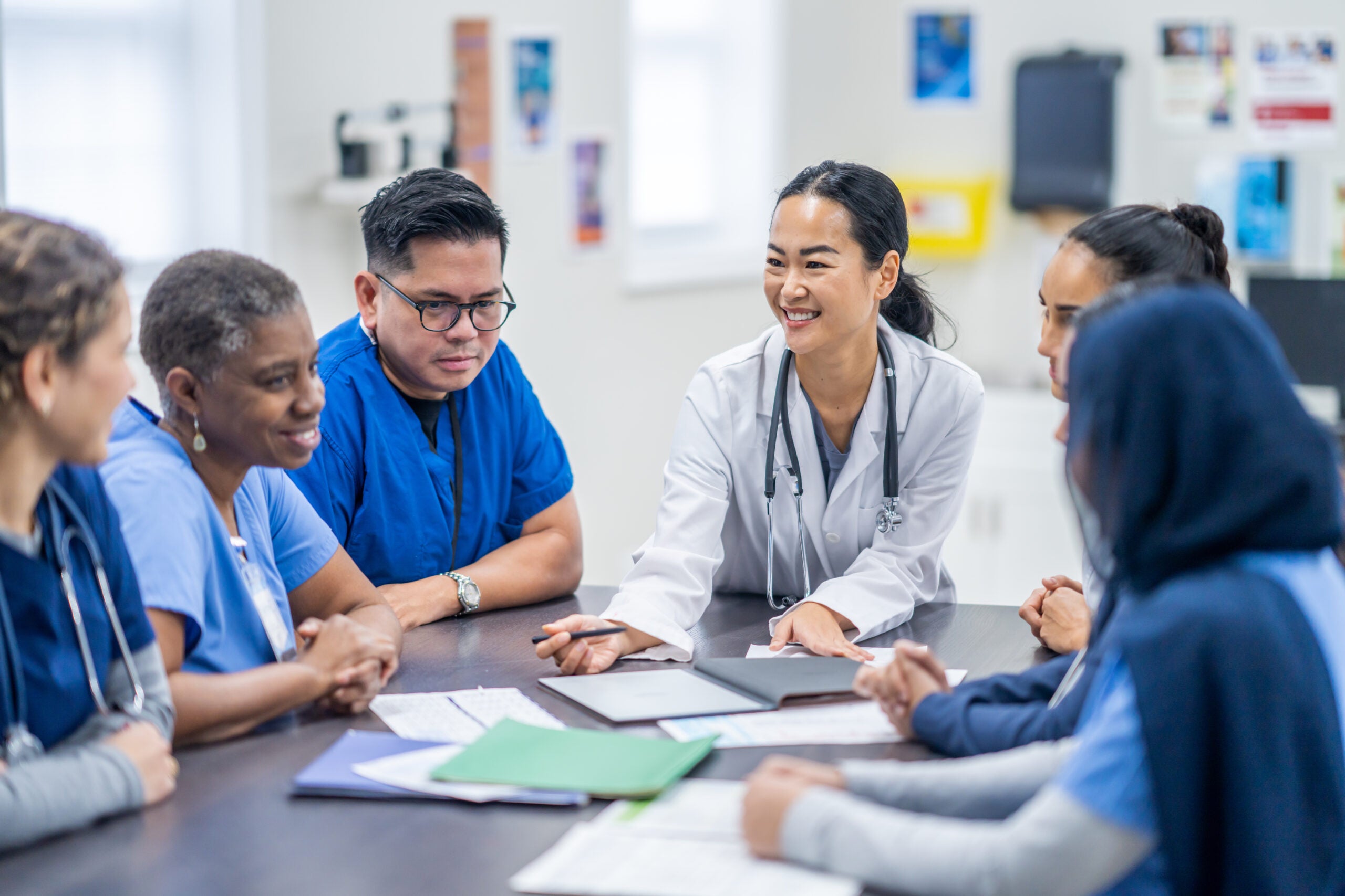 A small group of doctors and nurses gather around a meeting table to discuss patient cases. They are each dressed professionally and have various, files, notes and papers scattered between them as they reference them throughout the meeting.