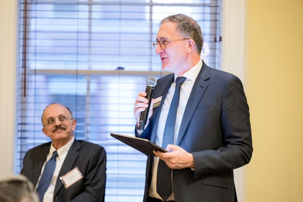 Dean Andrea Baccarelli, standing, speaks into a microphone held in his right hand, with a portfolio in his left hand. Wafaie Fawzi, seated in the background, looks on.