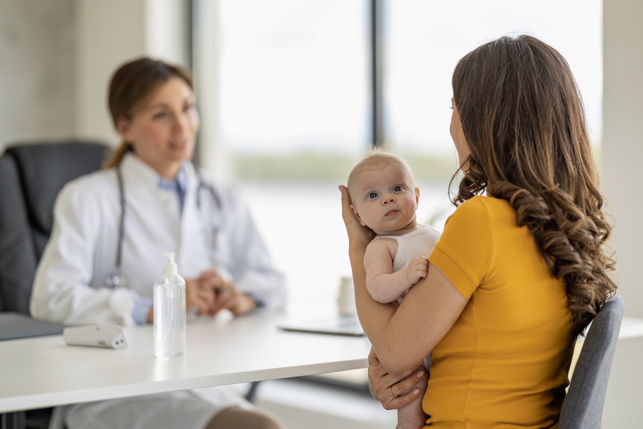 A mother holds her baby sitting in a chair across from a doctor. Mother faces doctor, baby faces the camera, in focus. Doctor is out of focus.