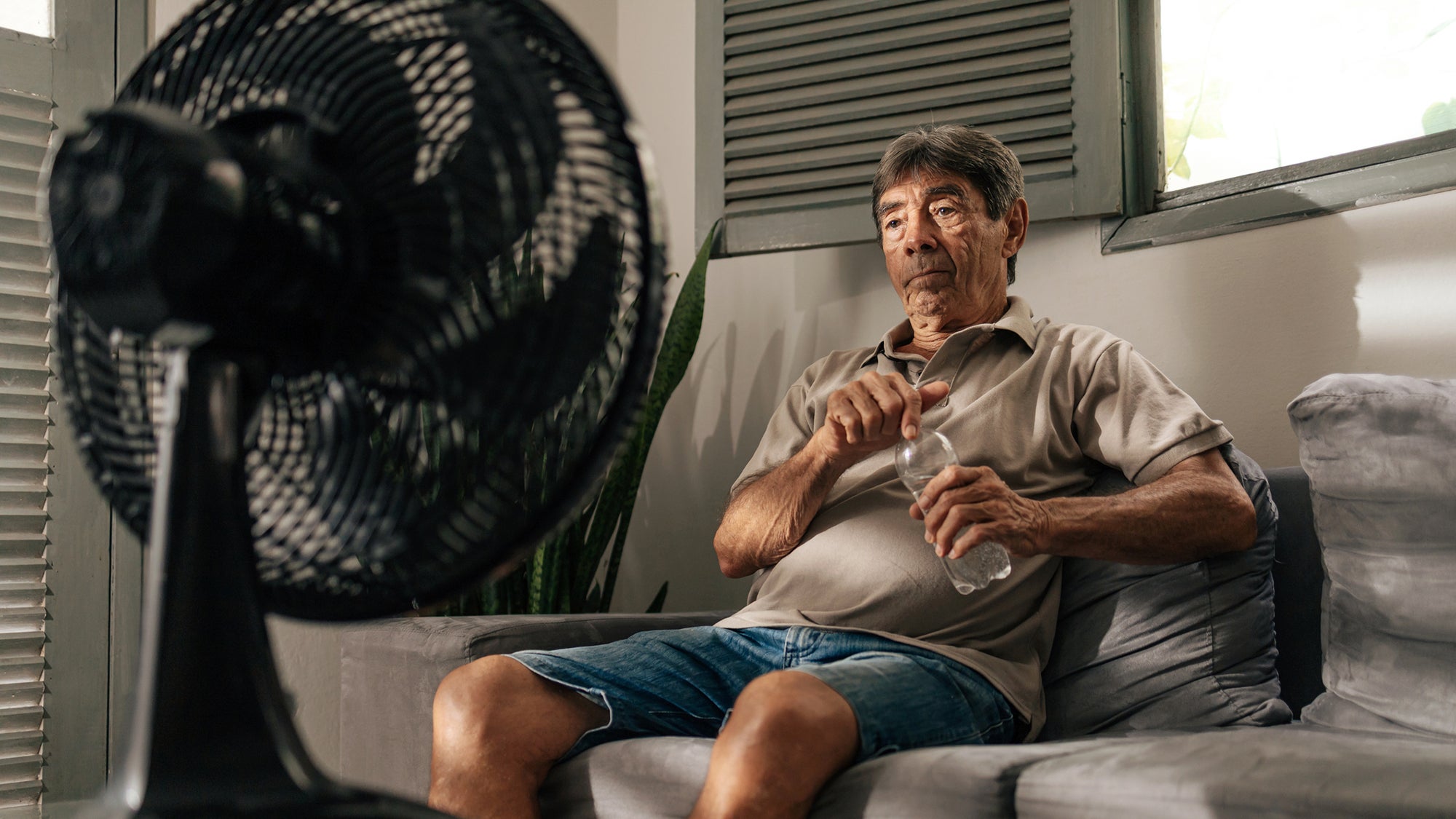 Senior man sits on a sofa at home using an electric fan to cool down on a hot day, holding a water bottle and resting