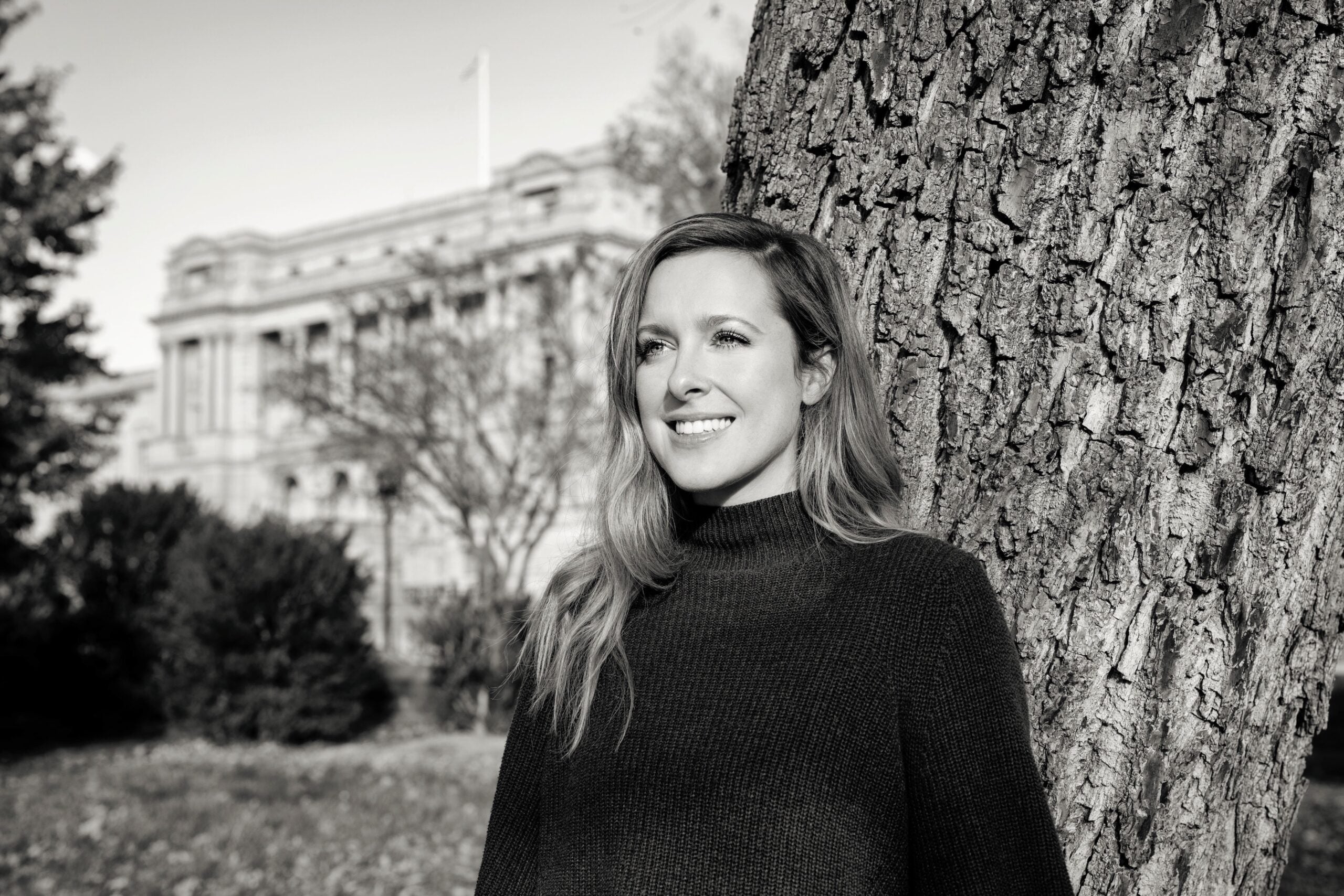 Maggie Penman is featured from the waist up in a black sweater with a tree behind her