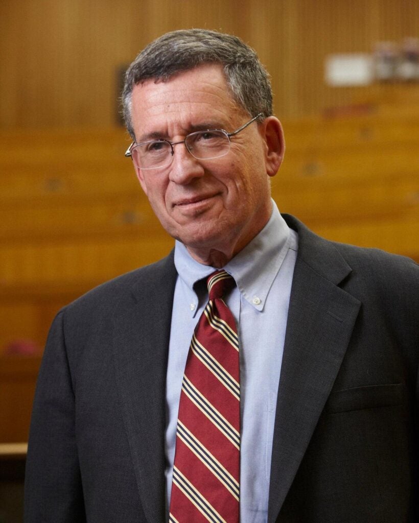 Portrait of Bob Blendon smiling, wearing a blazer and a red striped tie. In the background is a blurred classroom.