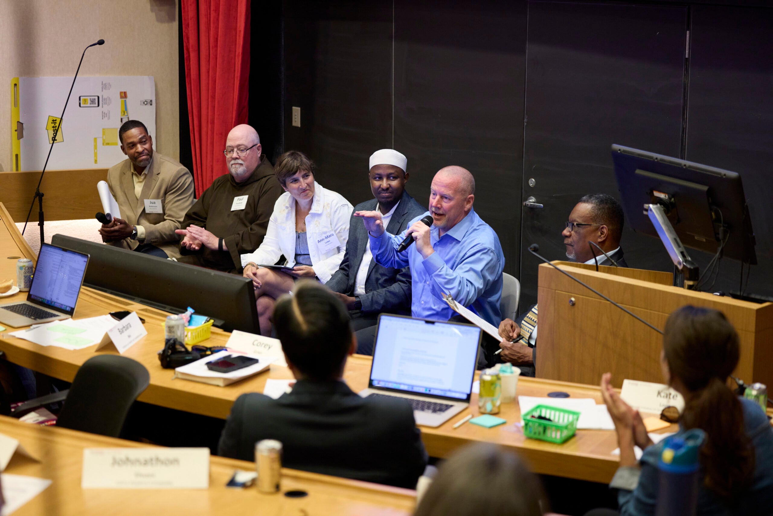 Six adults sit in a line at the front of a classroom. One is speaking into a microphone.
