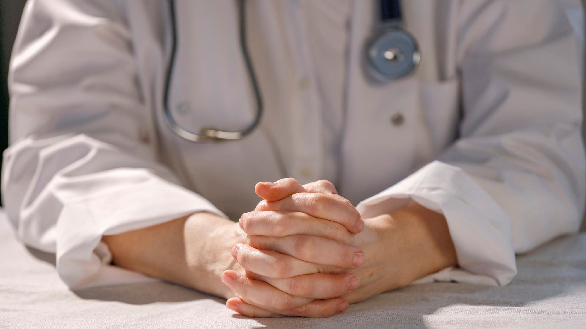 Doctor in white medical coat sitting at table with her hands folded.