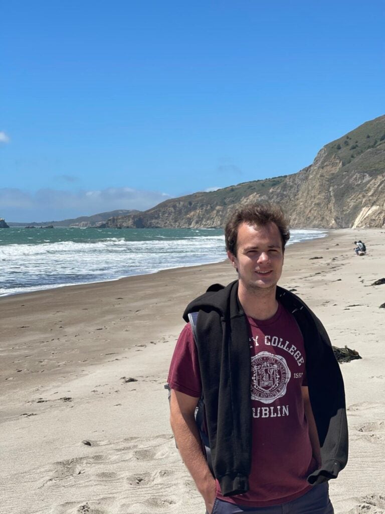 Eric standing on a beach, wearing a T-shirt, under a clear, blue sky.