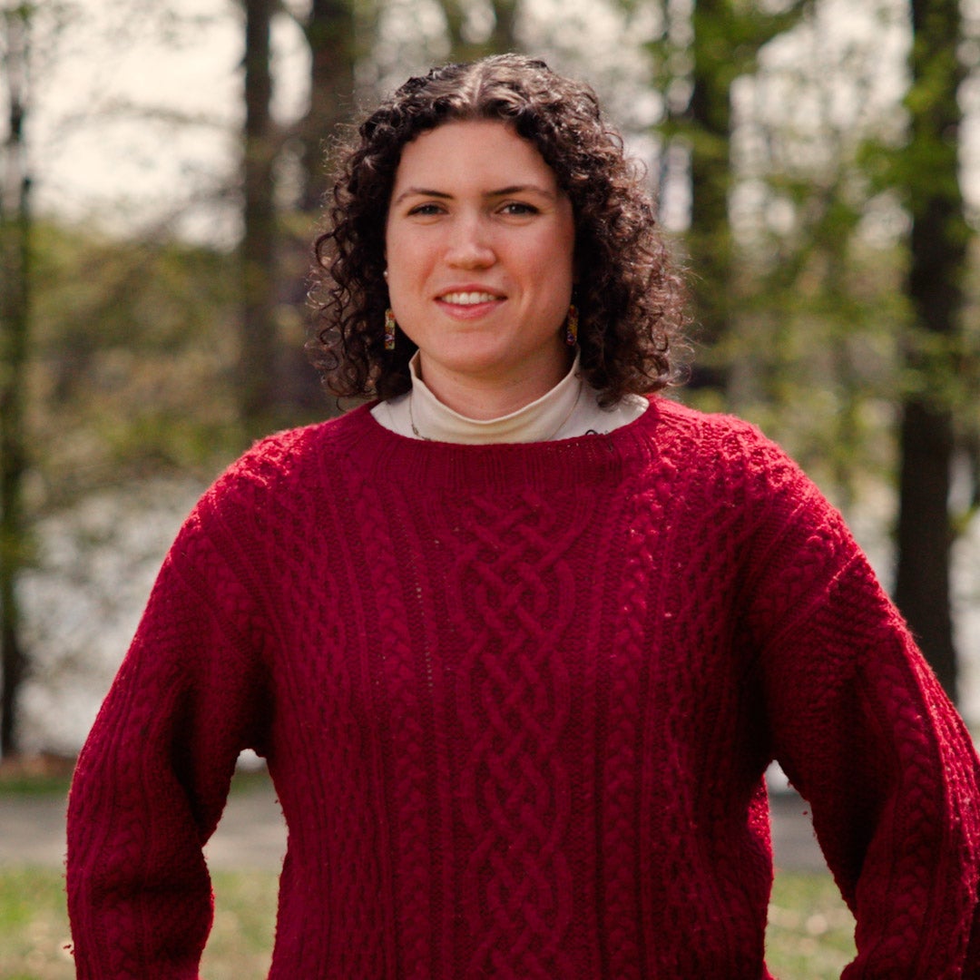 Outdoor, environmental headshot of Cora Cunningham. She wears a red cabled-sweater and stands near woods.