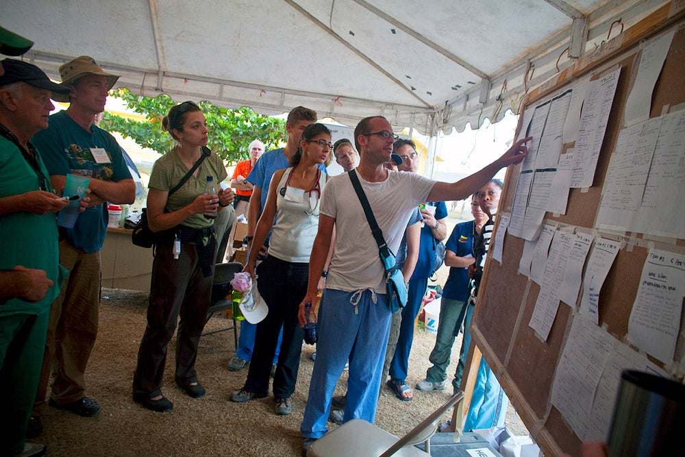 A public health professional speaks and points to papers posted on a bulletin board under a white tent. Other professionals are gathered, listening.