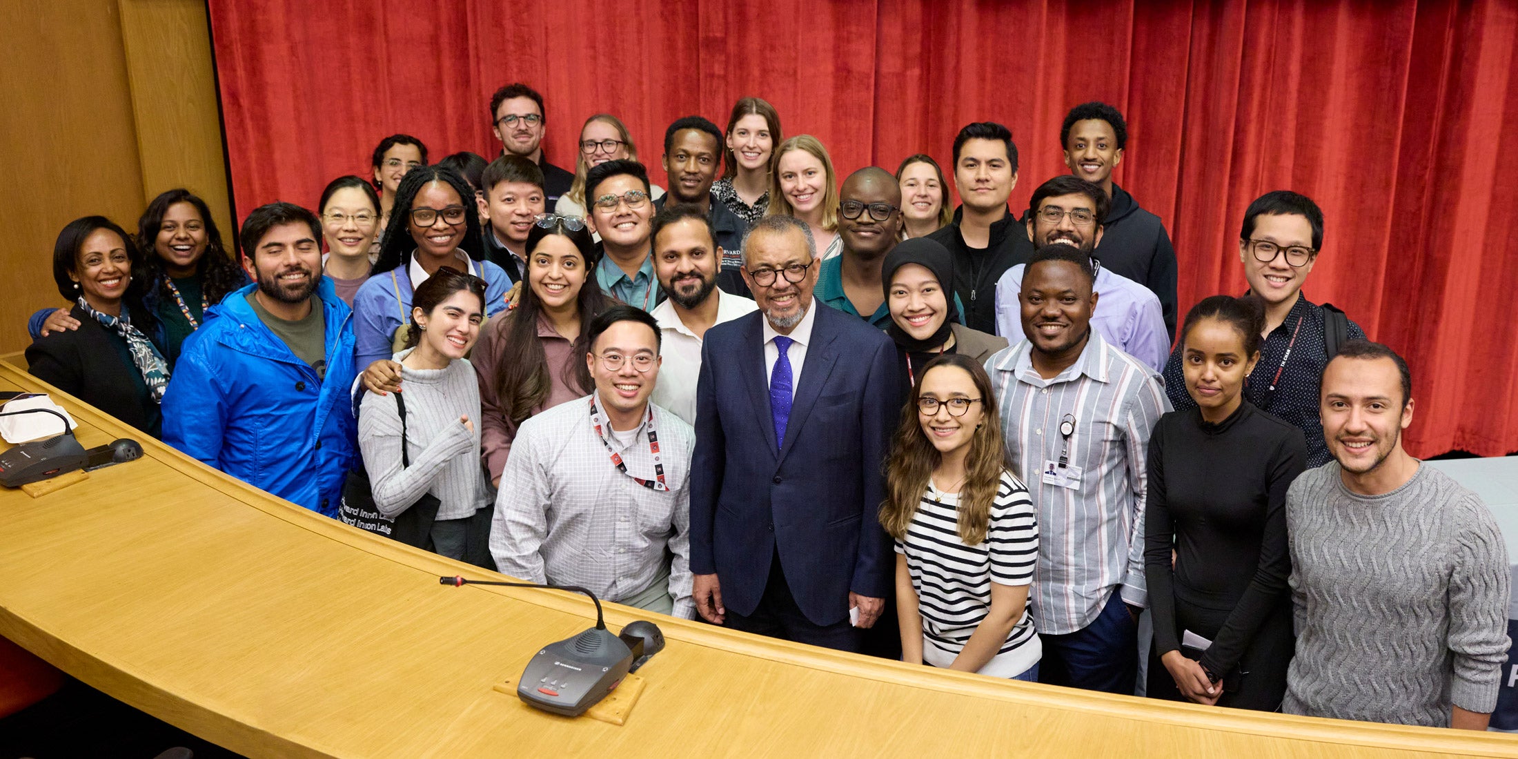 A large group of HSPH students pose for a photo with Tedros Adhanom Ghebreyesus in front of a red curtain in a lecture hall.