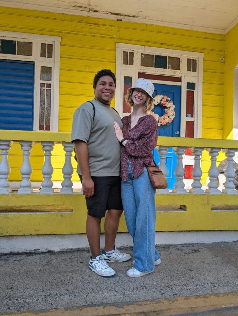 Madeline and her husband in Puerto Rico standing together outside a yellow home.