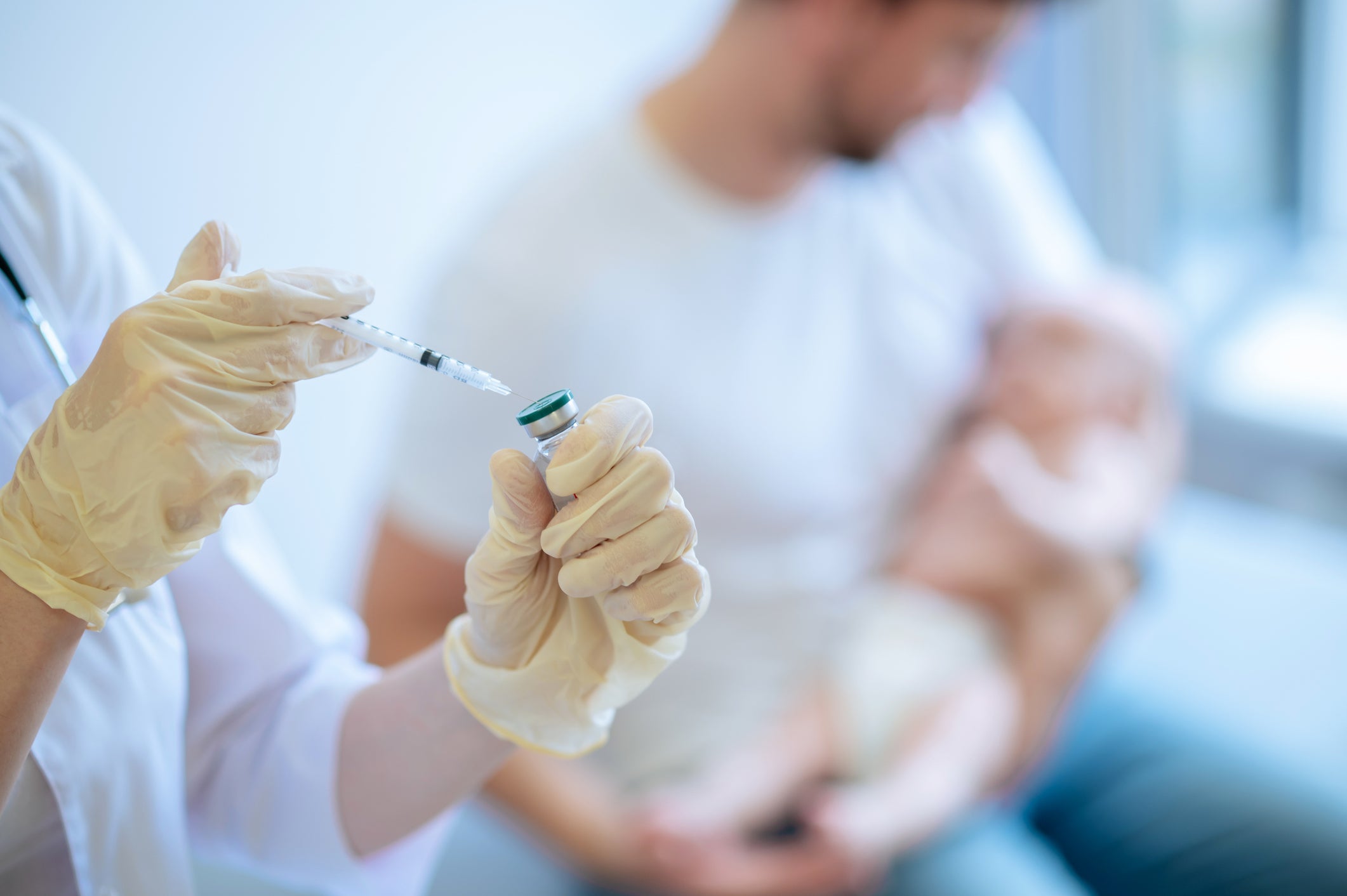 Cropped photo of a doctor in sterile gloves drawing up the medicine from the ampoule into the disposable syringe. A father holds a baby in the background, out of focus.