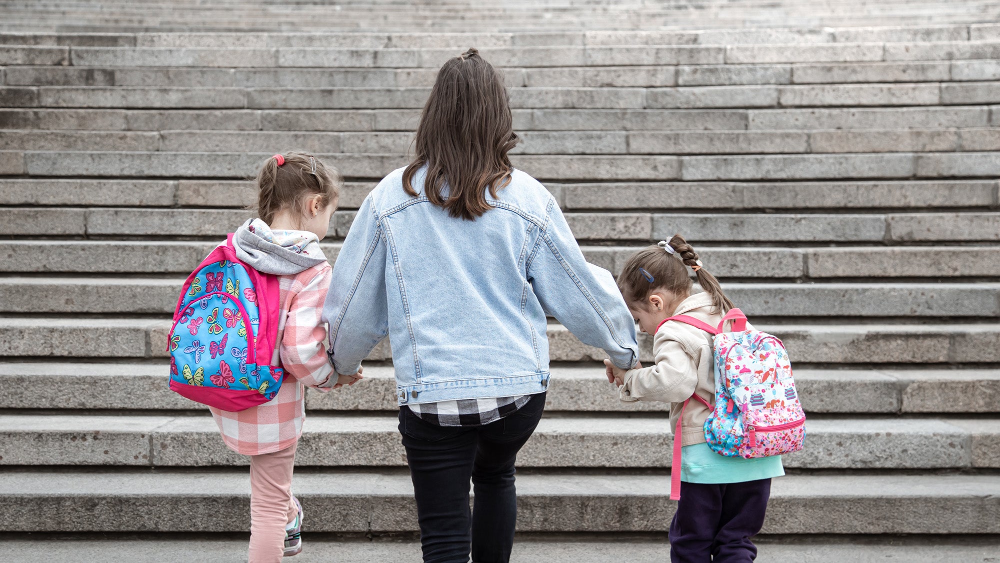 Mother and two daughters with backpacks, viewed from rear, walk hand in hand up a set of steps.