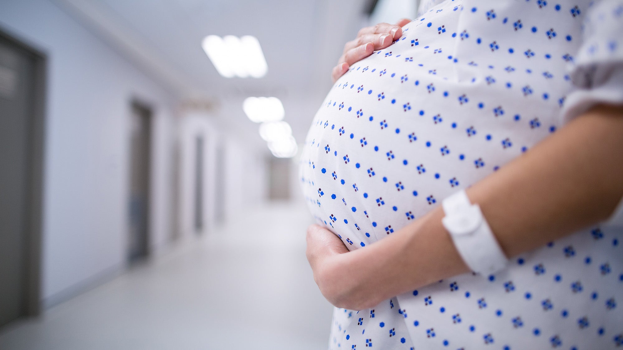 Pregnant woman in hospital gown, seen in mid-section, standing in hospital hallway.