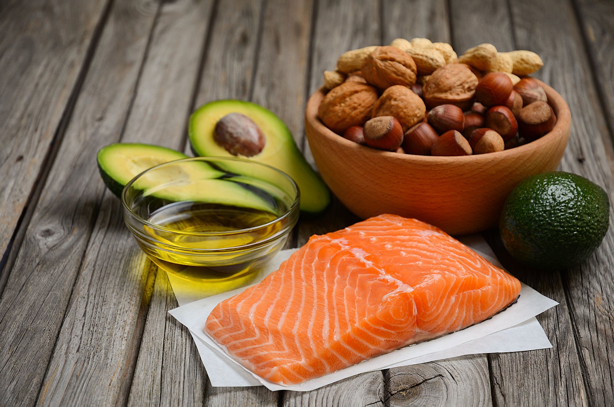A piece of raw salmon, avocados, and a bowl od nuts sit on a wooden table.