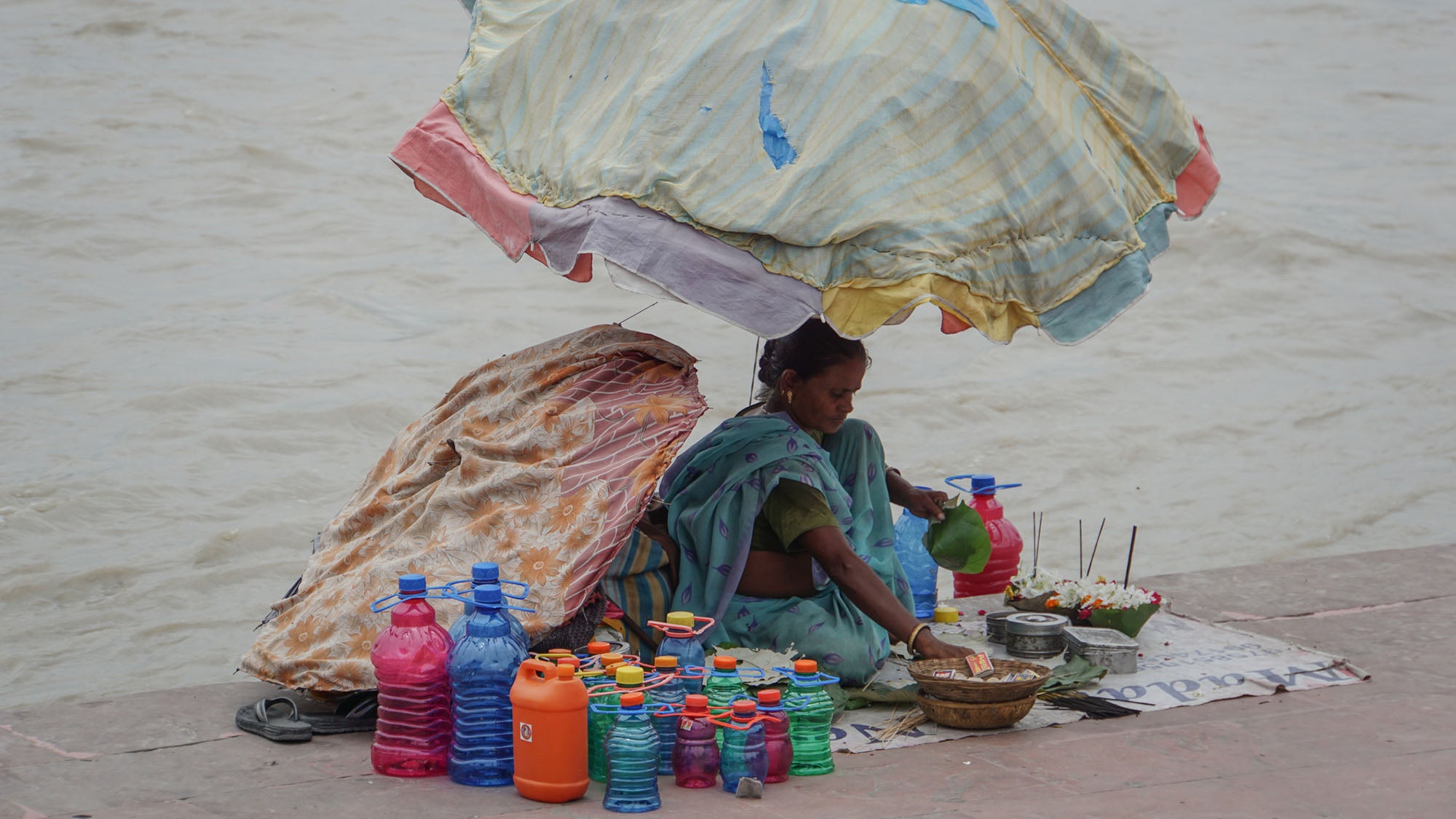 A woman in a blue sari sits under a makeshift striped umbrella, arranging items for sale on a white mat next to the muddy water of the Ganges River. Several colorful plastic bottles, metal bowls, small green leaf bowls with flowers, and burning incense sticks are displayed on the mat.