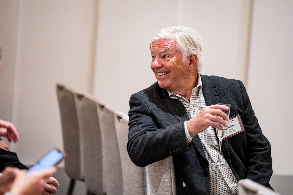 William Rom, holding a champagne glass and smiling, is turned around in his front-row seat to listen to someone out of frame. 