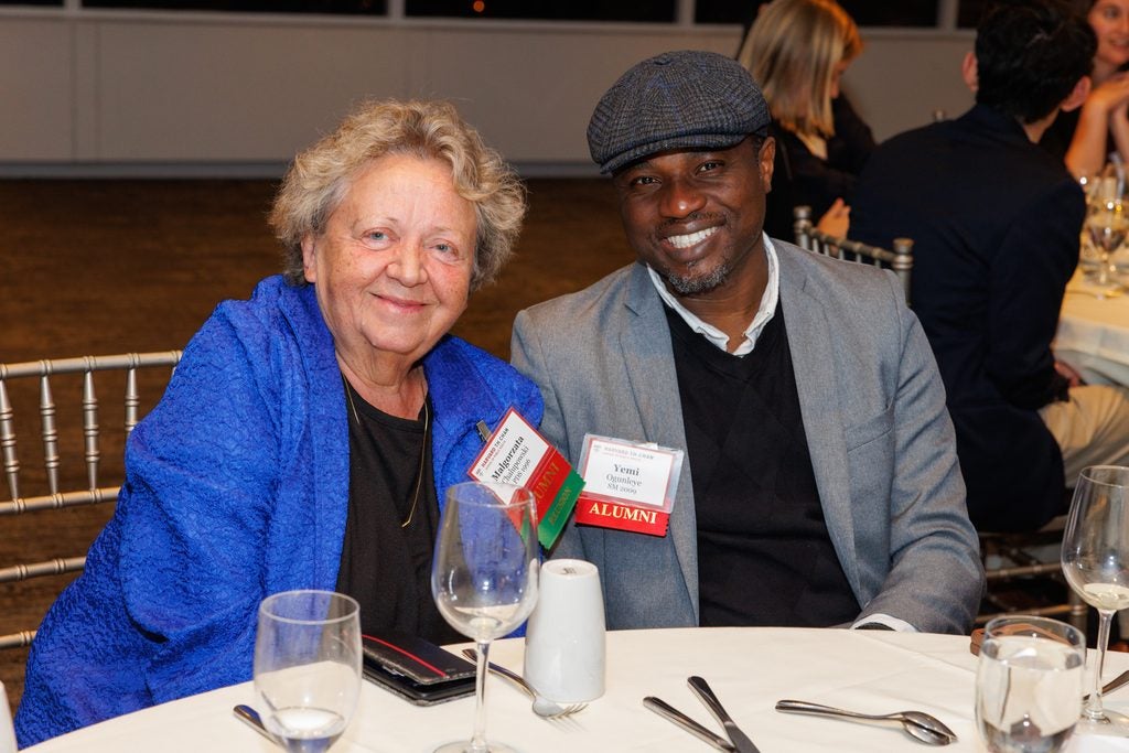 Two alumni lean together at a table, one wearing a newsboy hat.
