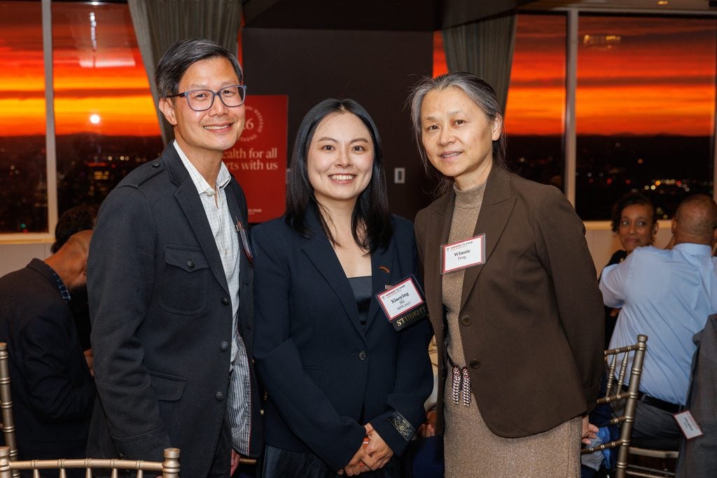 A student poses with two attendees in front of a dramatic red and orange sunset. 