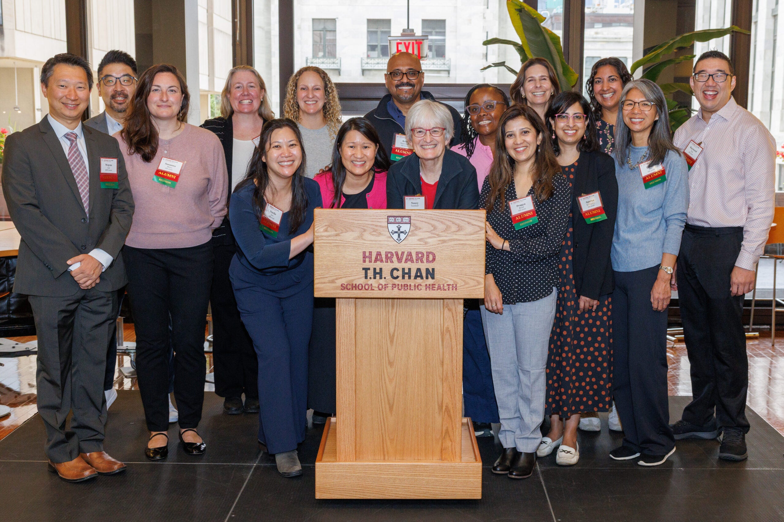 A group of fifteen alumni celebrating their reunion pose with Nancy Turnbull around the Harvard Chan School podium.
