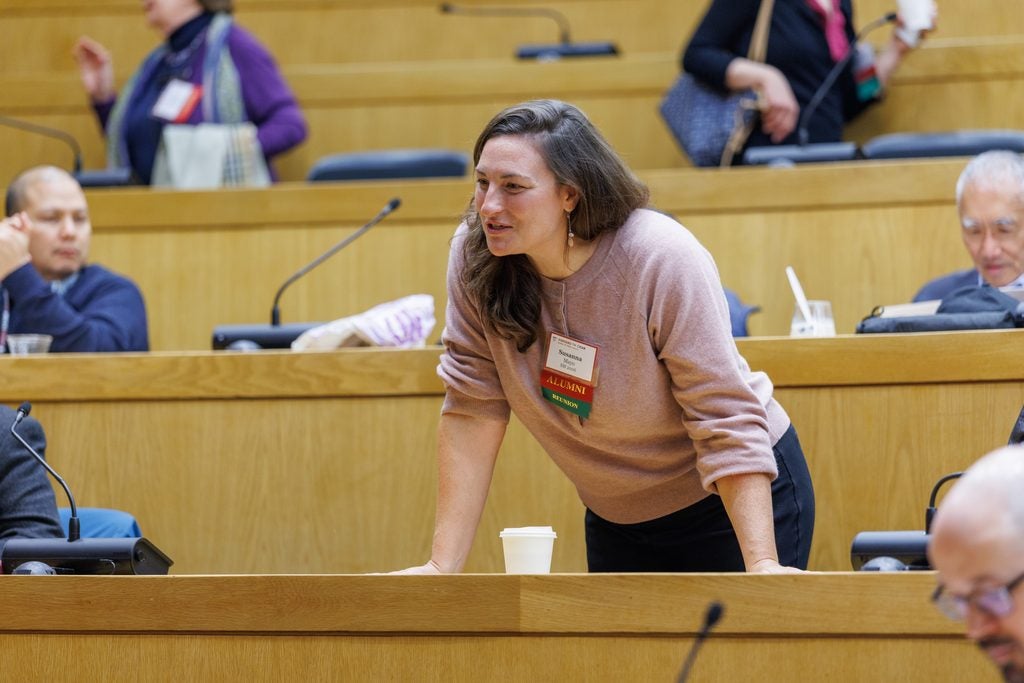 An alumna stands leaning forward on the table to speak from her seat in the lecture hall. 