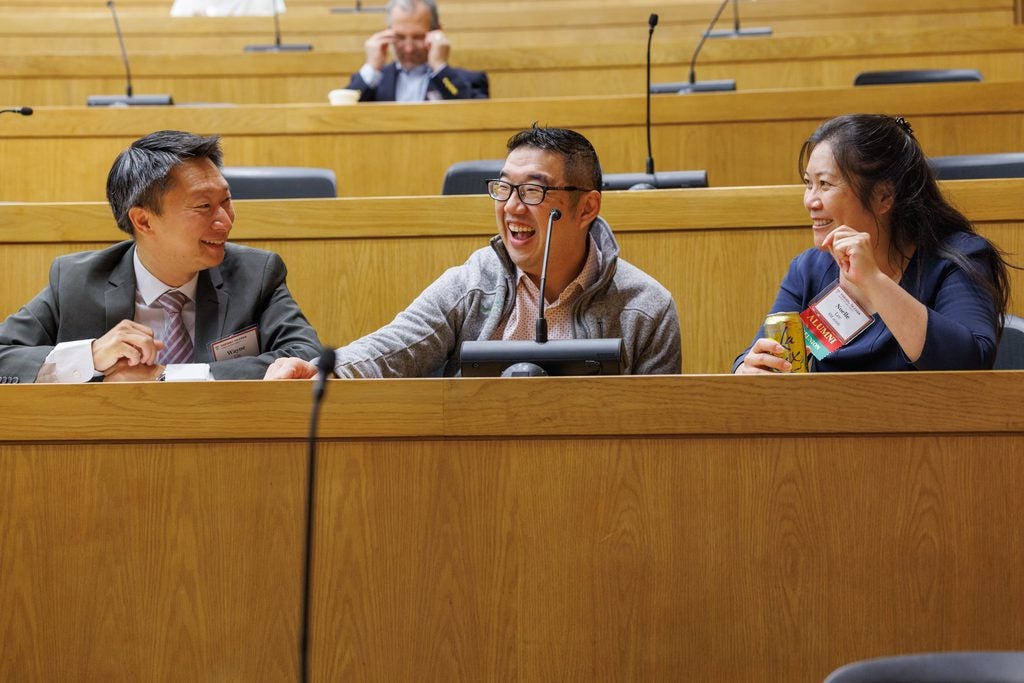 A group of three alumni laugh heartily in their lecture hall seats. 